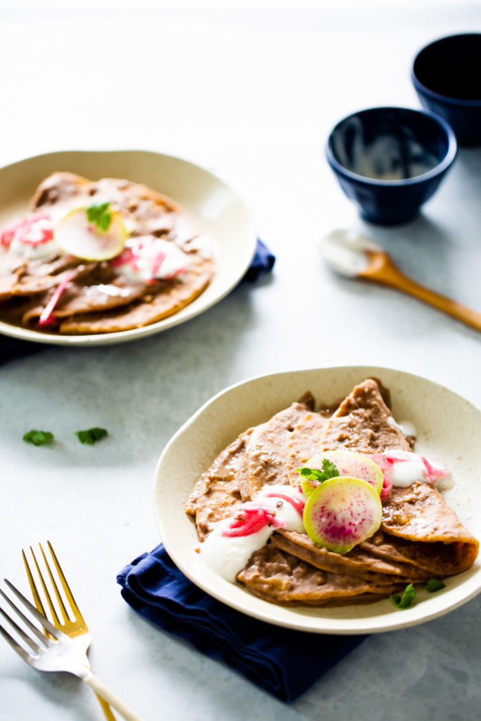 Enfrijoladas served on a plate finished with vegan crema, pickled onions and radishes.