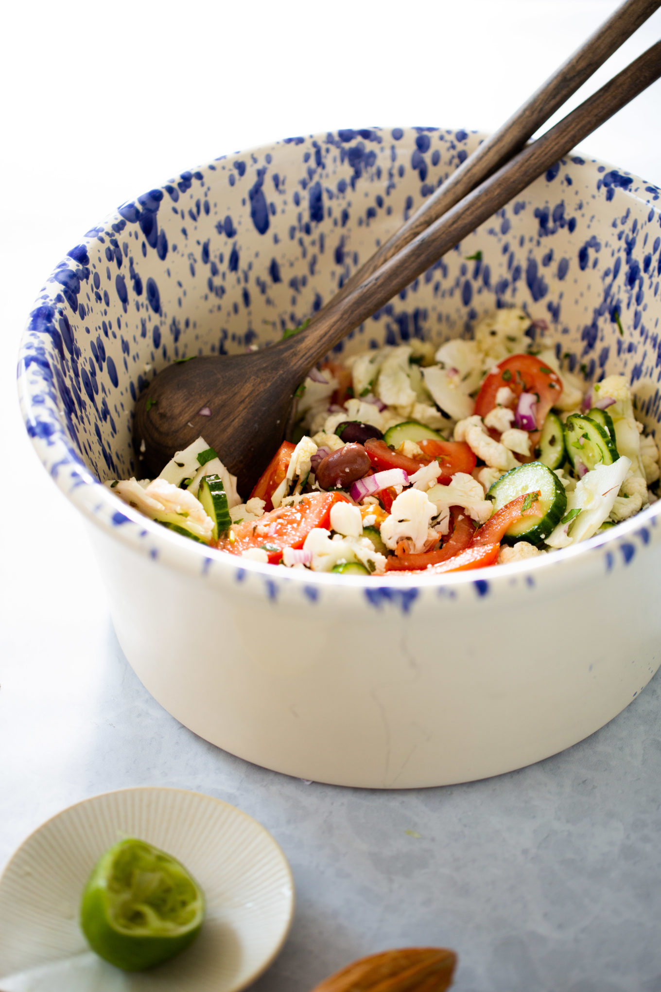 A large white bowl with blue speckles holds a fresh Mediterranean cauliflower salad of sliced cucumbers, tomatoes, red onion, olives, and crumbled feta, mixed with wooden salad servers. A squeezed lime half rests on a small plate nearby. The scene is bright and inviting.