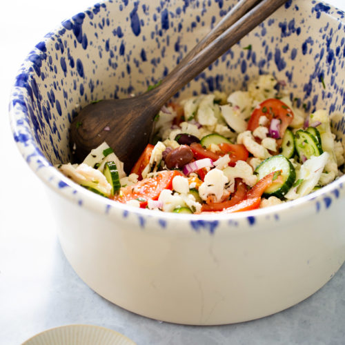 A large white bowl with blue speckles holds a fresh Mediterranean cauliflower salad of sliced cucumbers, tomatoes, red onion, olives, and crumbled feta, mixed with wooden salad servers. A squeezed lime half rests on a small plate nearby. The scene is bright and inviting.