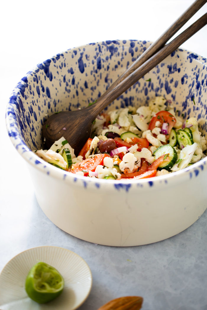 A large white bowl with blue speckles holds a fresh Mediterranean cauliflower salad of sliced cucumbers, tomatoes, red onion, olives, and crumbled feta, mixed with wooden salad servers. A squeezed lime half rests on a small plate nearby. The scene is bright and inviting.