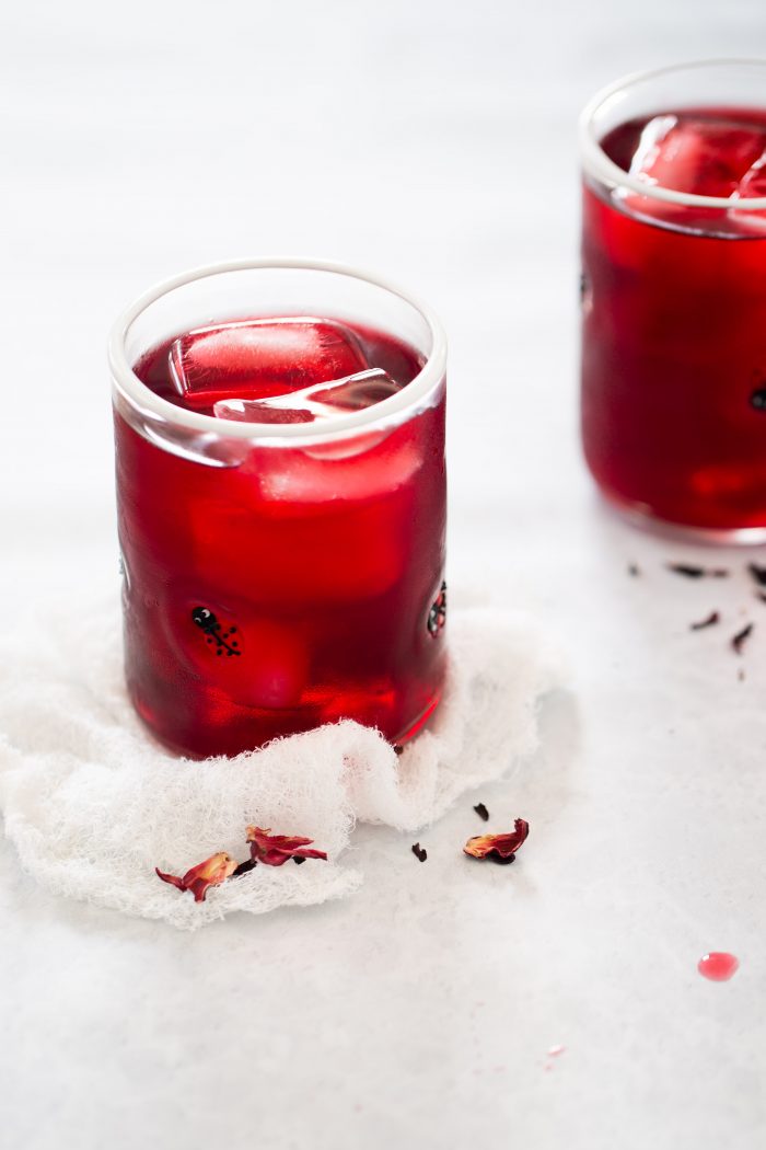Primer plano de un vaso lleno de una bebida roja y cubitos de hielo, colocado sobre una tela de color claro. Otro vaso similar está ligeramente desenfocado en el fondo. Hay pétalos de hibisco secos esparcidos alrededor de los vasos, lo que sugiere una bebida de agua de Jamaica. El fondo es una superficie gris clara.