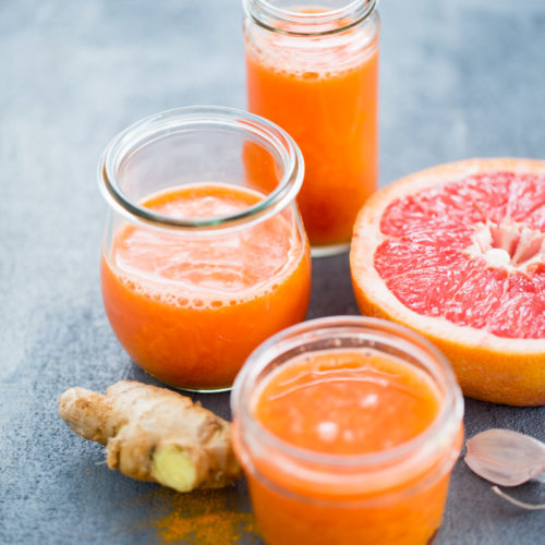 Tres frascos de pomelo fresco y jengibre sobre una superficie gris texturizada, con un pomelo partido por la mitad y un trozo de jengibre cerca. Destaca el vibrante color naranja del jugo.