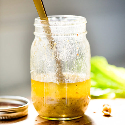 A glass mason jar filled with yellow simple vinaigrette, with seasoning settled at the bottom and a spoon inside. The jar sits on a wooden surface, metal lid beside it, and blurred lettuce leaves in the background as sunlight streams in from the left.