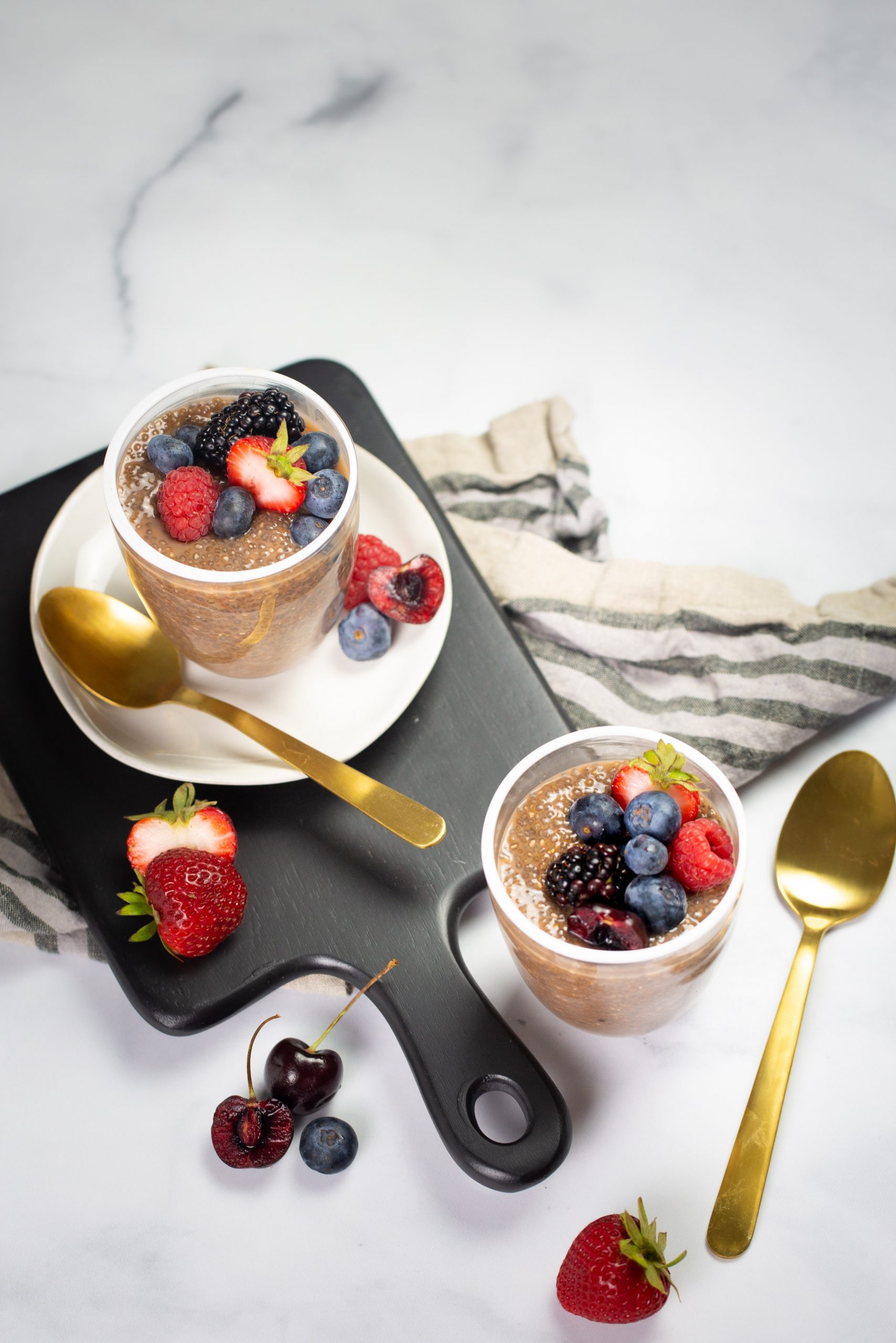 Dos tazas de pudín de chocolate y chía cubierto con moras, frambuesas y arándanos descansan elegantemente sobre un plato blanco y una tabla rectangular negra. Cucharas doradas acompañan cada taza, mientras que fresas y cerezas adornan la tabla. Un mantel a rayas agrega un toque de encanto contra la superficie de mármol blanco.