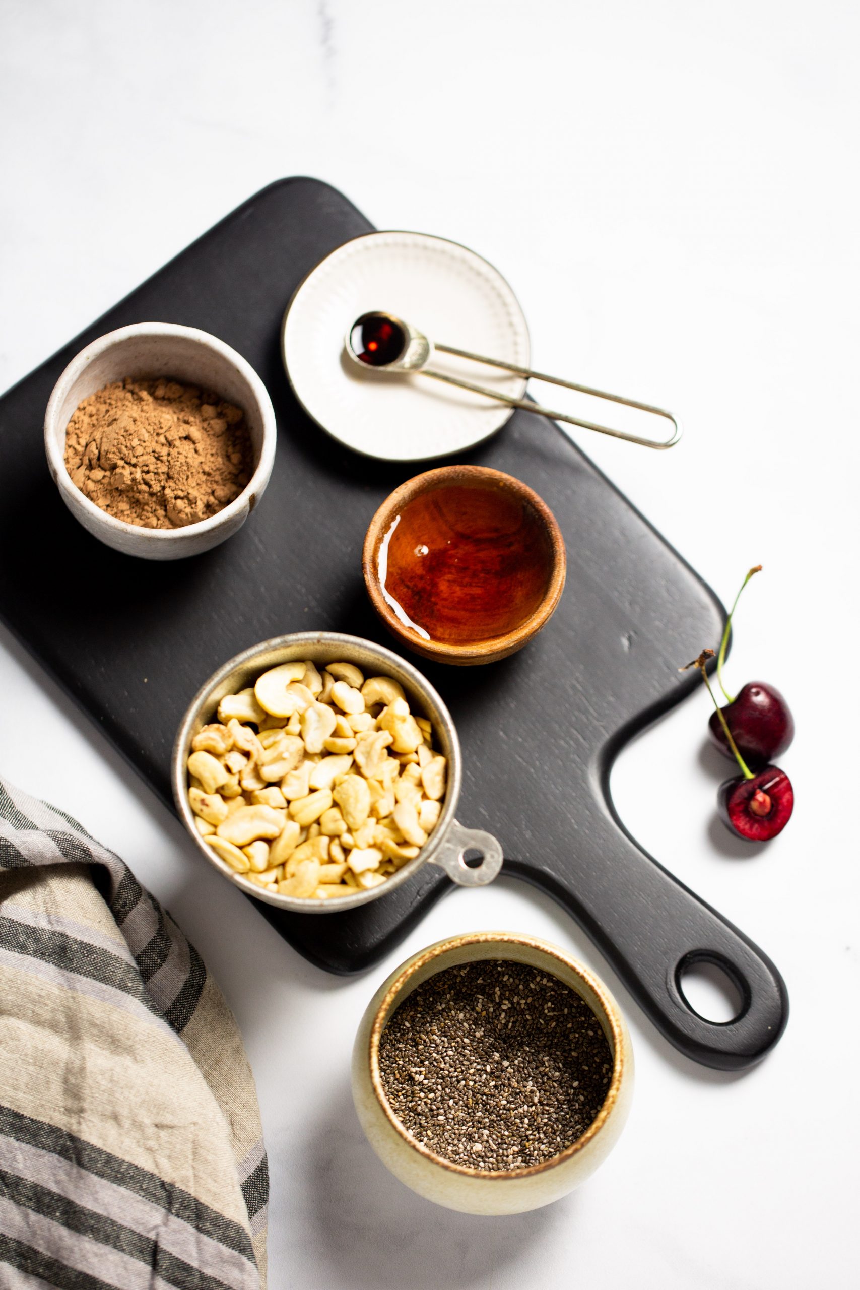 Una tabla de cortar negra muestra los ingredientes para preparar pudín de chocolate y chía: cacao en polvo en un bol blanco, anacardos en una taza medidora de metal, miel o almíbar en un bol de madera y semillas de chía en un plato de cerámica. Un plato blanco pequeño sostiene una cuchara con líquido oscuro. Dos cerezas y un mantel a rayas añaden encanto al conjunto.