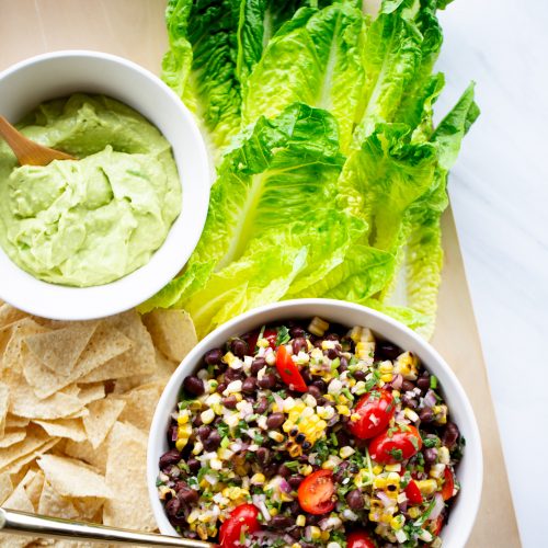 A wooden board showcases a bowl of black bean salad mixed with corn, tomatoes, and herbs, flanked by whole romaine lettuce leaves. Nearby, enjoy a bowl of creamy guacamole and crispy tortilla chips. The salad glimmers with flavors akin to pico de gallo with black beans, a spoon ready for serving.
