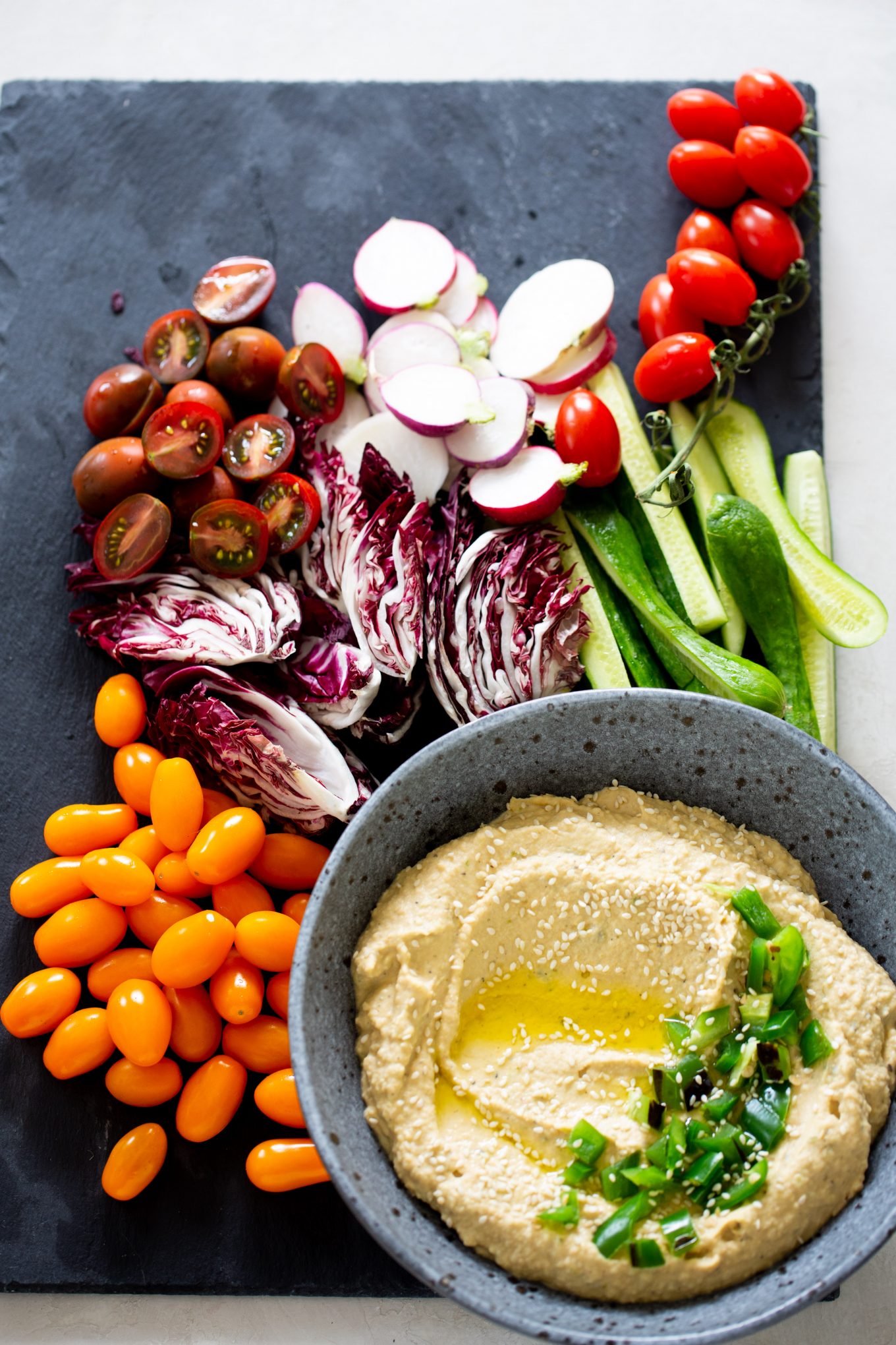 A bowl of jalapeño hummus with chopped green onions and sesame seeds on top is placed on a slate. Surrounding it are sliced radishes, cherry tomatoes, halved purple radicchio, cucumber spears, and whole orange grape tomatoes arranged neatly for serving.
