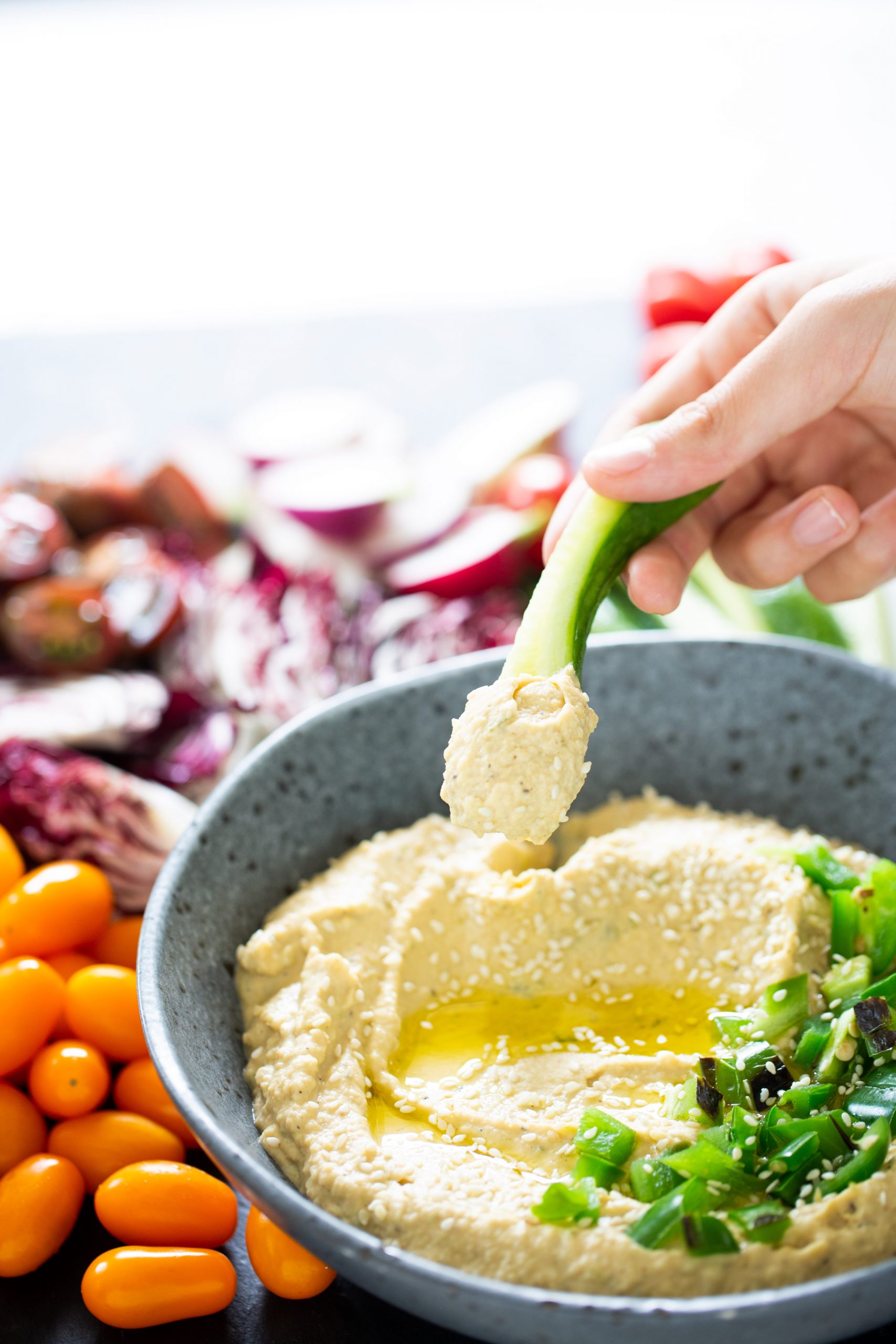 A hand dips a green onion into a bowl of creamy jalapeño hummus topped with sesame seeds and herbs. The hummus is drizzled with olive oil. Surrounding the bowl are bright cherry tomatoes and slices of radish and cabbage, all placed on a dark surface.