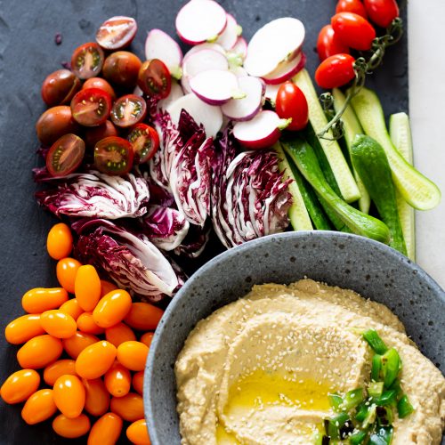A bowl of jalapeño hummus with chopped green onions and sesame seeds on top is placed on a slate. Surrounding it are sliced radishes, cherry tomatoes, halved purple radicchio, cucumber spears, and whole orange grape tomatoes arranged neatly for serving.