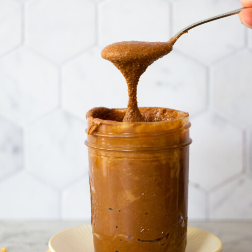 A spoon lifts a dollop of gooey, dark chocolate peanut butter from a full mason jar set on a small cream-colored plate. Peanut pieces are scattered on the gray countertop. The background features a white hexagonal tile pattern.