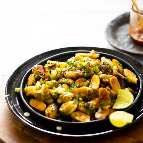 A black plate of sweet and spicy Brussels sprouts garnished with herbs and green onions sits on a wooden serving board. Lime wedges are on the side. A small jar with a spoon is in the background, slightly blurred. The setting is bright and minimalistic.