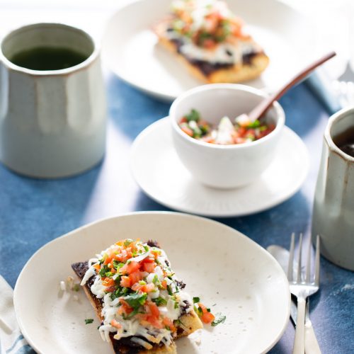 A plate of molletes features a crispy tortilla topped with refried beans, melted cheese, diced tomatoes, and chopped herbs. A small bowl of salsa sits on the blue table beside it, with a fork and knife nearby. Two ceramic mugs and another plate of similar delights blur in the background.