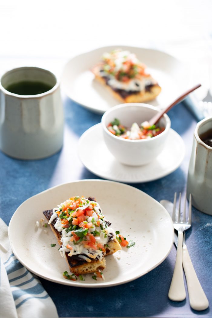 A plate of molletes features a crispy tortilla topped with refried beans, melted cheese, diced tomatoes, and chopped herbs. A small bowl of salsa sits on the blue table beside it, with a fork and knife nearby. Two ceramic mugs and another plate of similar delights blur in the background.