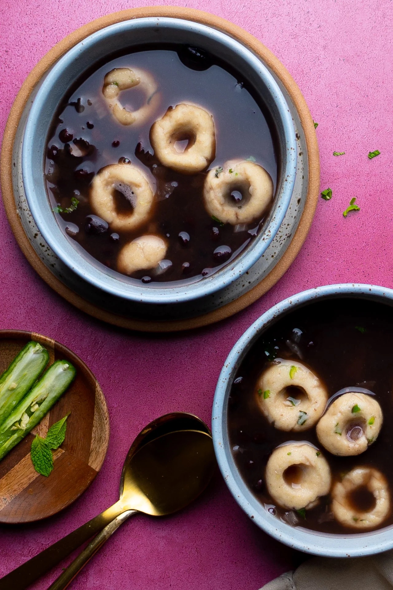 A top view of two bowls of vegan soup with dumplings, garnished with herbs, on a pink surface. A wooden plate with three cucumber slices and a mint leaf, a gold spoon, and a beige napkin sit next to the bowls. The soup is dark with light, round dumplings floating in it—perfect for your vegan Mexican recipes collection.