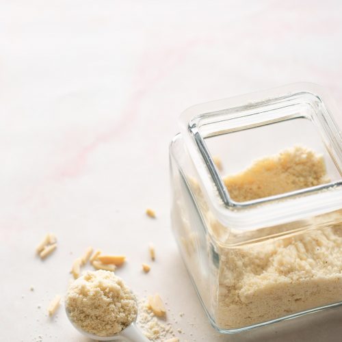 A glass jar filled with almond flour sits on a light surface. Some almond flour is spilled beside the jar, ready to be used for almond flour cookies, while a white ceramic tablespoon and a few slivered almonds rest nearby.