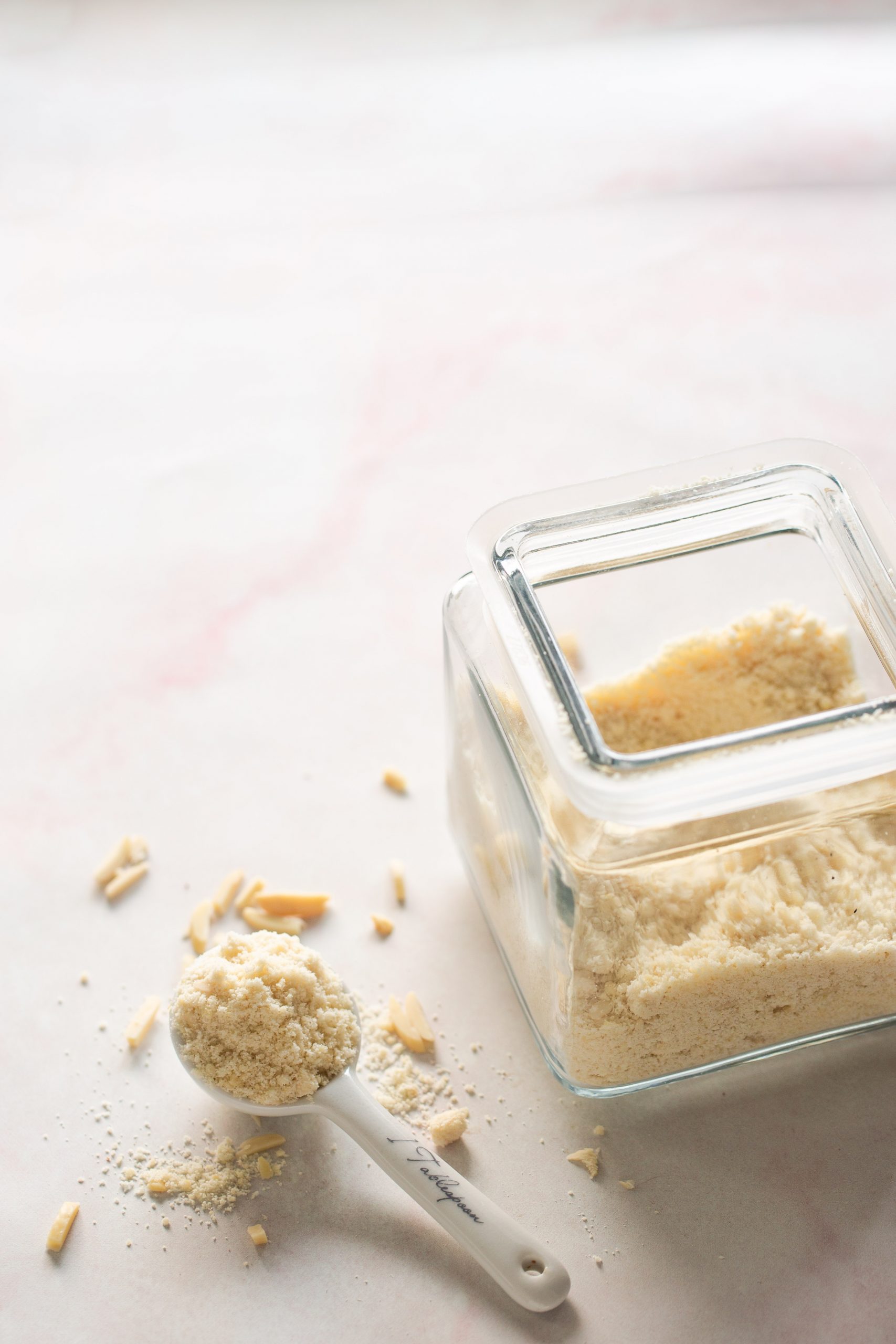 A glass jar filled with almond flour sits on a light surface. Some almond flour is spilled beside the jar, ready to be used for almond flour cookies, while a white ceramic tablespoon and a few slivered almonds rest nearby.