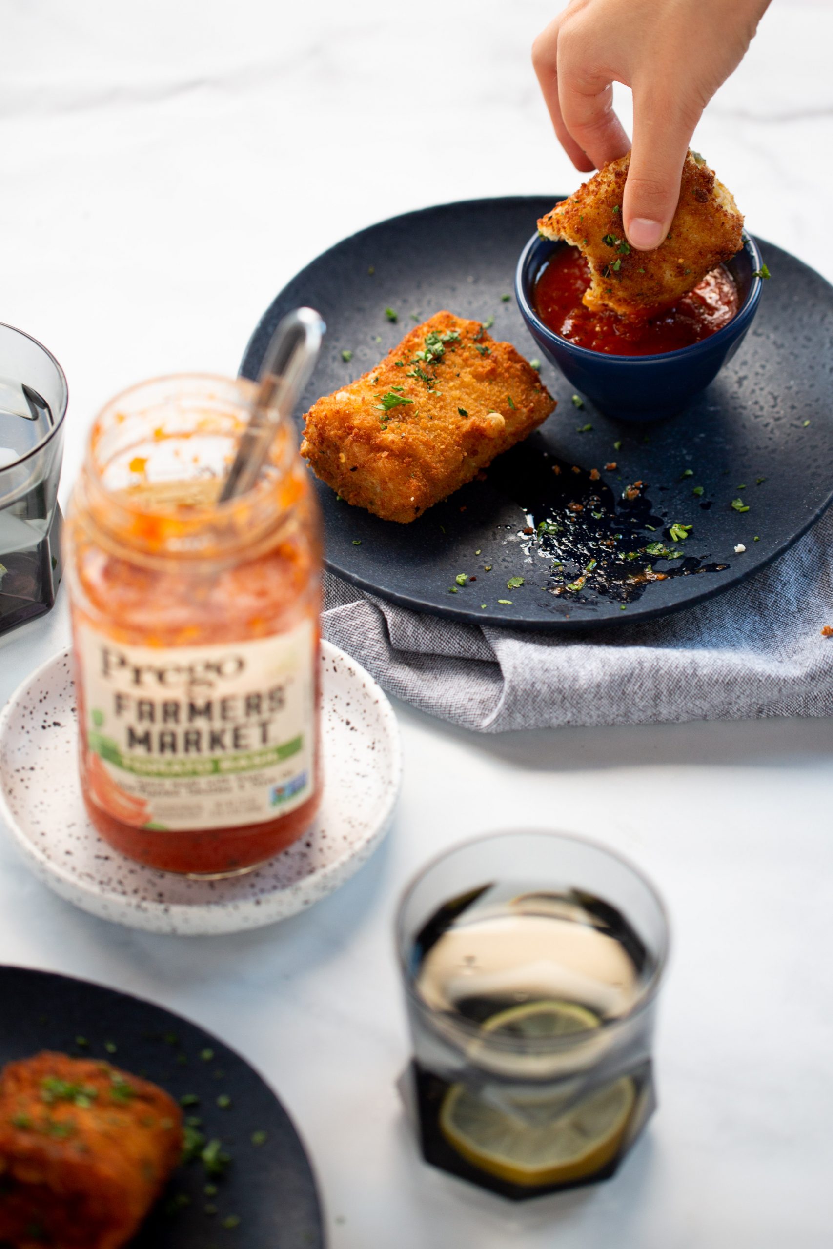 A hand dips a fried lasagna roll-up into a small bowl of red marinara sauce on a dark plate. Beside it is another crispy roll-up, garnished with herbs, along with pasta sauce, a soda with ice, and utensils on a white surface.