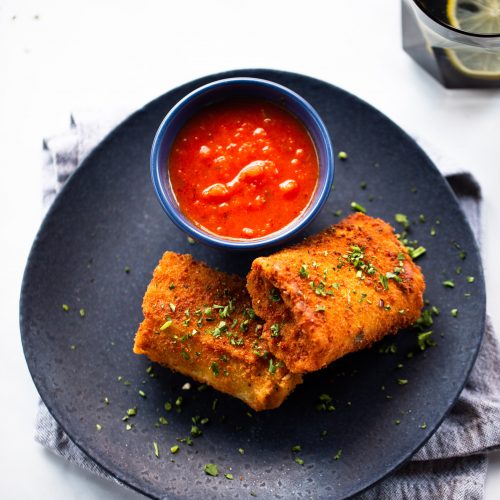A dark plate holds two deep-fried lasagna rolls garnished with herbs, next to a blue bowl of red dipping sauce. The plate rests on a folded gray cloth, with a glass of water and a lemon slice visible in the top right corner.