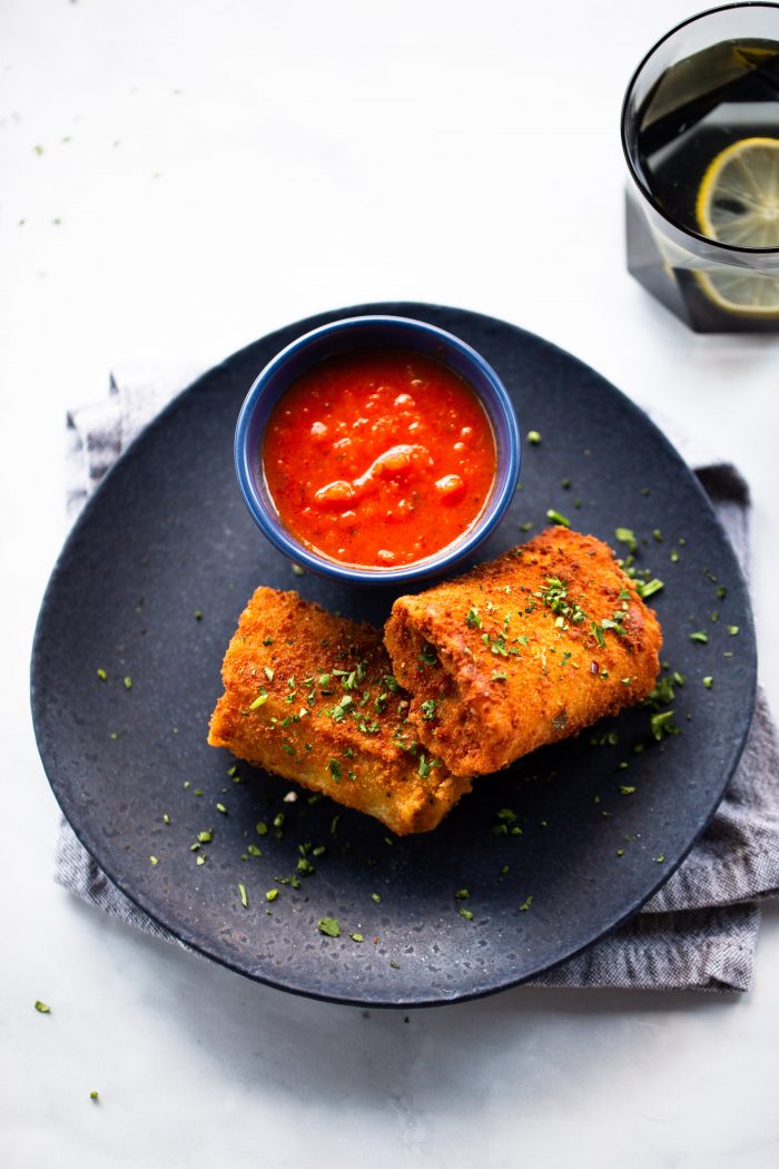 A dark plate holds two deep-fried lasagna rolls garnished with herbs, next to a blue bowl of red dipping sauce. The plate rests on a folded gray cloth, with a glass of water and a lemon slice visible in the top right corner.