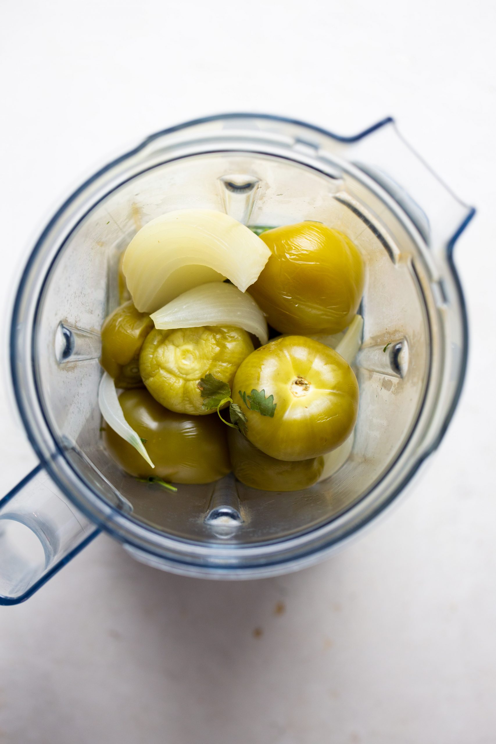 Overhead view of a blender containing whole cooked tomatillos, large onion chunks, and cilantro on a white surface, ready to be blended for a green sauce or salsa perfect for no-bake enchiladas.