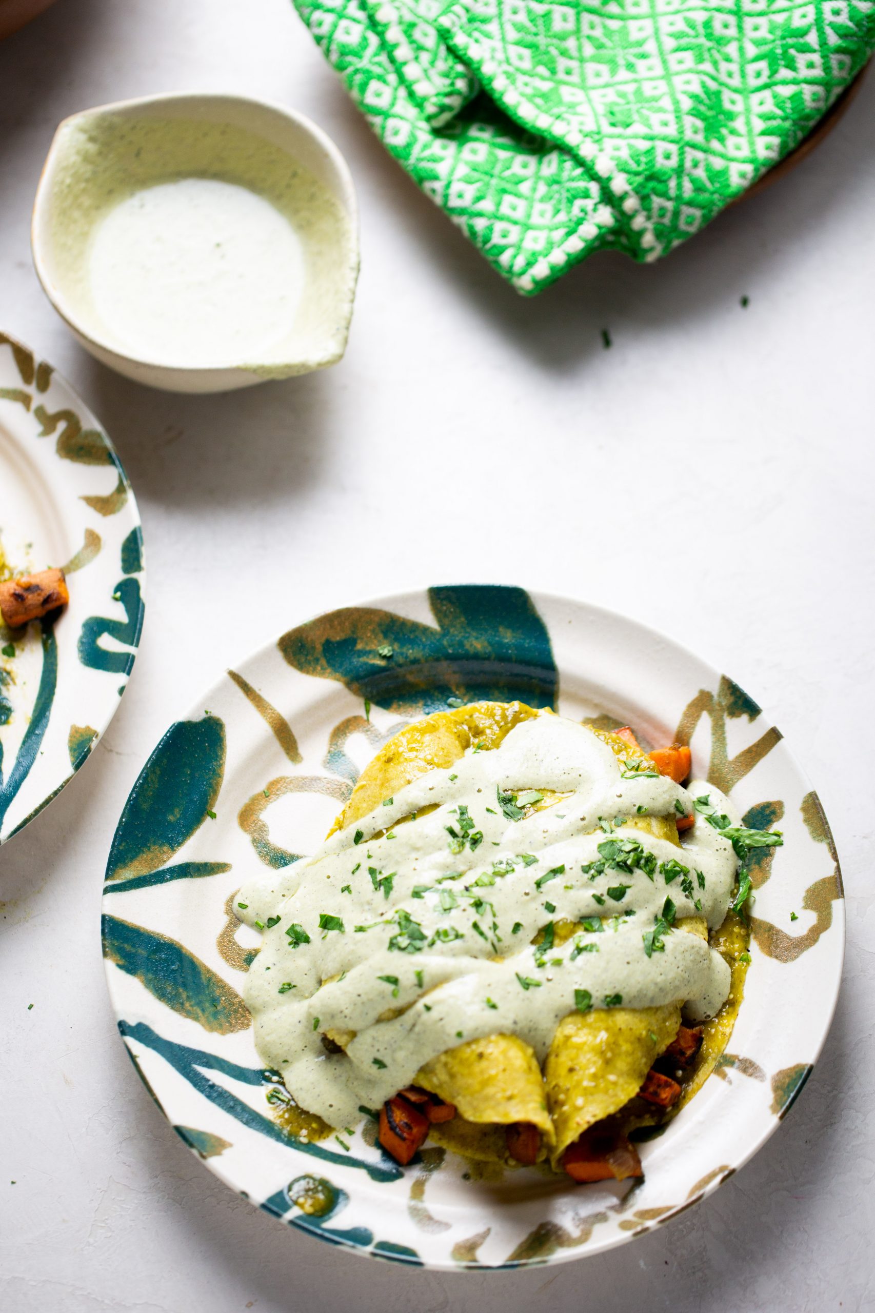 A plate of no-bake enchiladas is topped with creamy green sauce and fresh herbs. A bowl of sauce and a green patterned cloth sit nearby, all arranged on a white surface with a multicolored plate featuring blue and gold patterns.