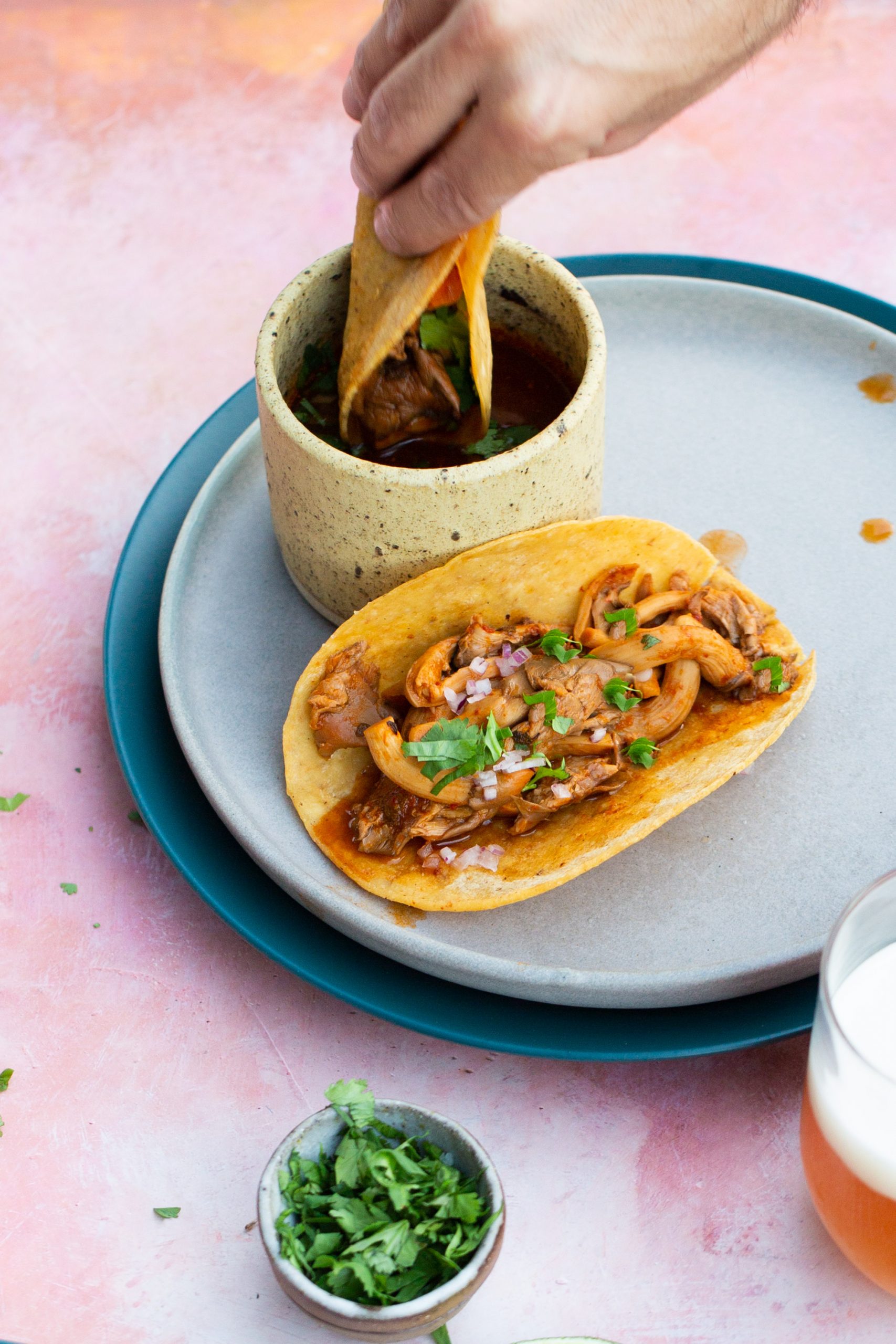 A hand dips a mushroom birria taco filled with shredded meat into a cup of consommé. Another taco topped with meat, chopped onions, and cilantro sits on a gray plate beside a small bowl of cilantro and a glass of beer on a pink surface.