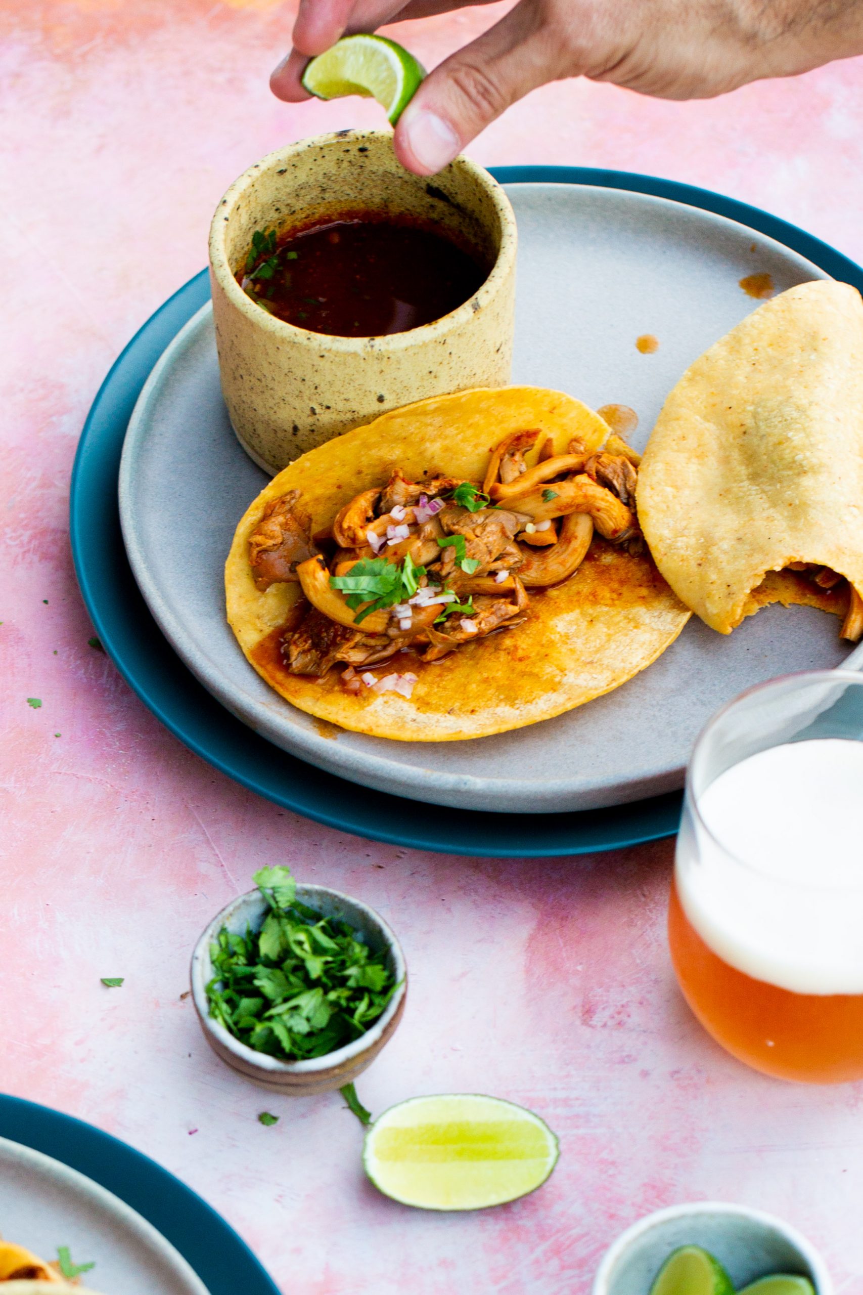 A hand squeezes a lime wedge over vegan birria tacos filled with shredded meat, onions, and cilantro on a gray plate, next to a cup of red salsa and another folded taco. Nearby are a glass of beer, chopped cilantro, and lime wedges on a pink surface.