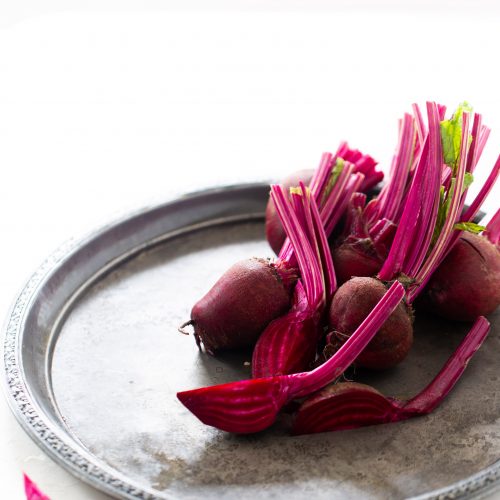 A silver tray holds several fresh beets with long, vibrant pink stems and leaves. One beet is sliced, revealing its deep red interior. The tray is placed on a white surface, and a small beet slice rests nearby, adding a touch of color contrast.