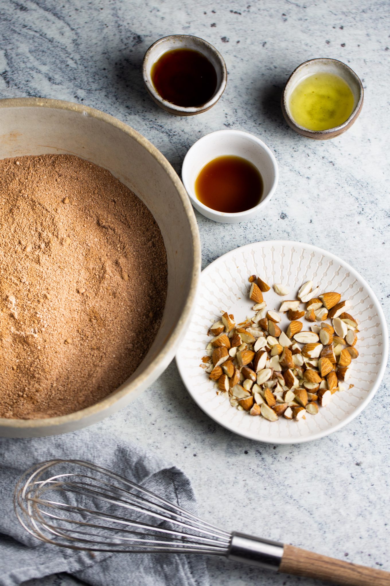 Bowl with dry ingredients and a plate of almonds on the side. Dishes with vanilla and oil.