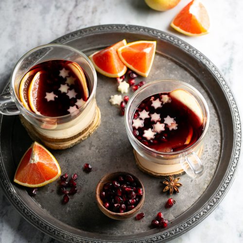 Two glass mugs of spiced mulled wine topped with star-shaped slices on a round tray. Garnished beautifully with orange slices, a small bowl of pomegranate seeds, cinnamon sticks, and star anise. Apples and orange segments grace the light gray marbled surface.