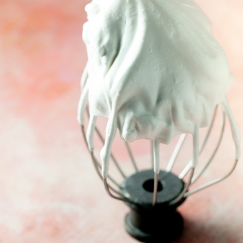 A metal whisk attachment stands upright on a pink surface, covered in glossy, soft peaks of white whipped aquafaba pavlova, highlighting its airy texture. The background is softly blurred, enhancing the focus on the vegan pavlova's smooth, fluffy consistency.