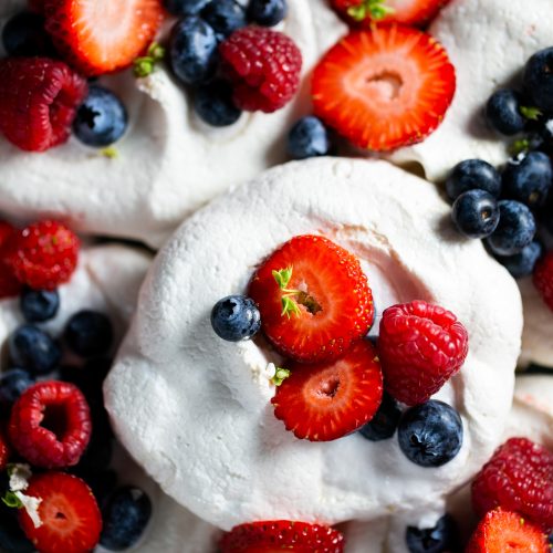 Close-up of vegan pavlova nests topped with fresh fruit. Each meringue is adorned with vibrant strawberries, blueberries, and raspberries. The meringues have a light, airy texture, contrasting with the colorful fruits. The composition highlights the freshness and natural colors.