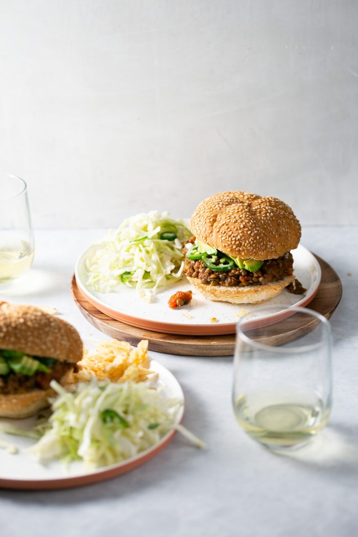 Two plates with sloppy joe-style sesame seed burgers, topped with lettuce and sliced cucumber, sit on a white surface. Each plate includes shredded cabbage salad, and two glasses of white wine are nearby against a plain, light-colored background.