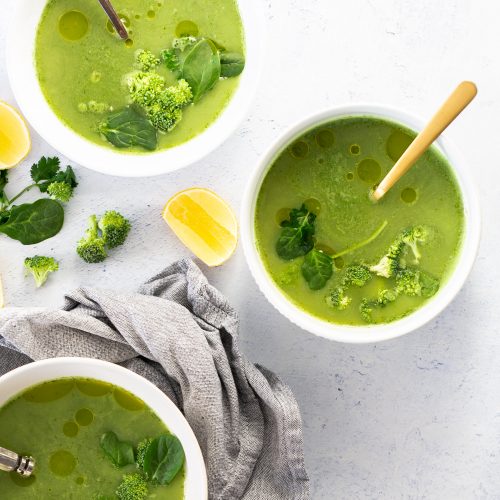 Three bowls of green soup garnished with leafy greens, broccoli florets, and herbs are arranged on a light surface. Spoons rest in each bowl. A gray cloth napkin, lemon wedges, and extra broccoli florets are scattered nearby, enhancing the fresh and healthy presentation.