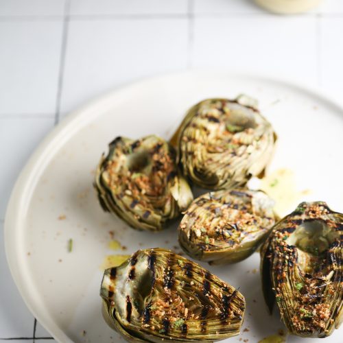 On a white tiled surface rests a white plate adorned with four grilled artichoke halves, seasoned with herbs and drizzled with olive oil. In the background, hinting at how to make grilled artichokes truly delicious, is a small jar filled with vibrant yellow sauce.