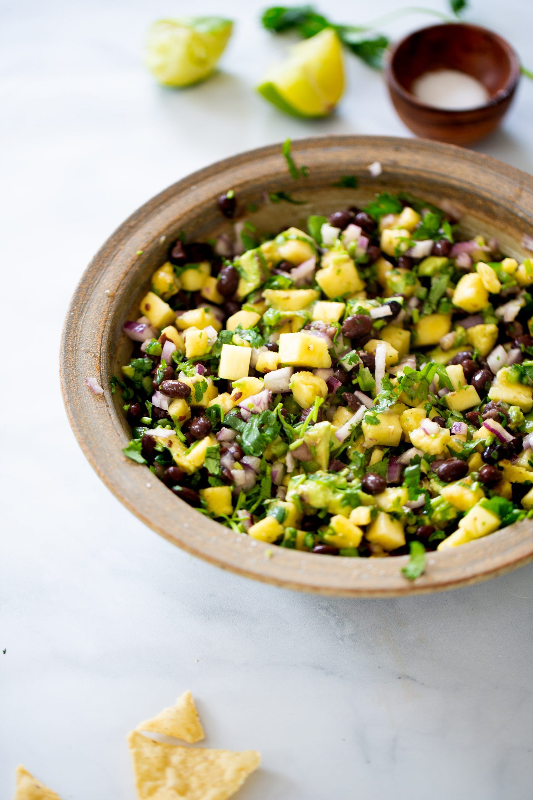 Pineapple Black Bean Salsa A rustic brown bowl filled with a colorful salad of diced mango, black beans, red onion, cilantro, and avocado—perfect as a topping for a Rice Bowl or served alongside Peach Salsa. Lime wedges, salt, and tortilla chips add the finishing touch on a white surface.