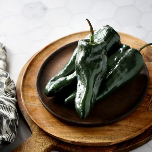 A quartet of dark green poblano peppers rests on a round wooden cutting board with a handle, set upon a hexagonal white tile surface. To the left, a patterned cloth peeks into view. The scene is bathed in soft, natural lighting.