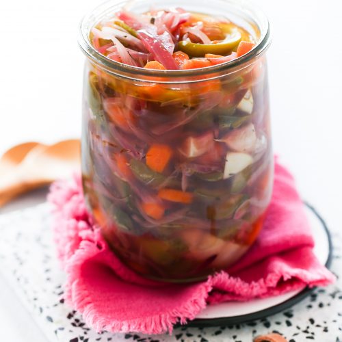 A glass jar filled with colorful pickled vegetables, including carrots, green beans, peppers, onions, and zesty Mexican pickled jalapenos. The jar sits on a pink napkin atop a speckled white plate with a brown leather loop, against a bright white background.