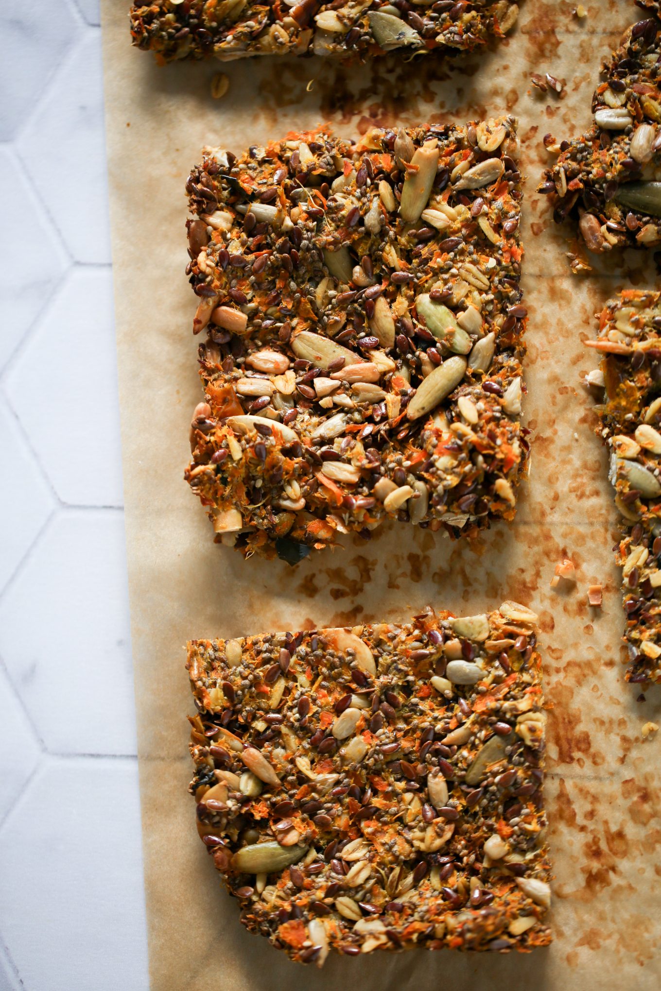 Three rectangular seed and nut bars, resembling tempting juice pulp crackers, rest on a parchment-lined surface. They boast visible pumpkin seeds, flaxseeds, and chopped nuts. Set against a light-colored countertop, their coarse, crunchy texture is highlighted in their close arrangement.