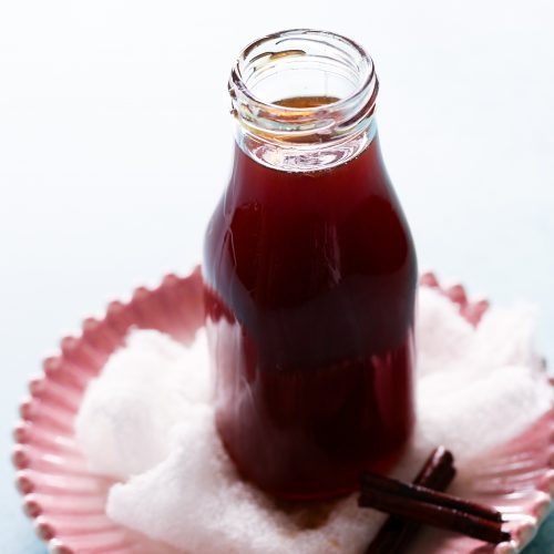 A glass bottle filled with dark liquid sits on a pink, scalloped dish, evoking curiosity about its contents—perhaps piloncillo syrup. Surrounding the bottle is a layer of white fabric or paper, with two cinnamon sticks placed beside it. The softly lit background creates a calm atmosphere.