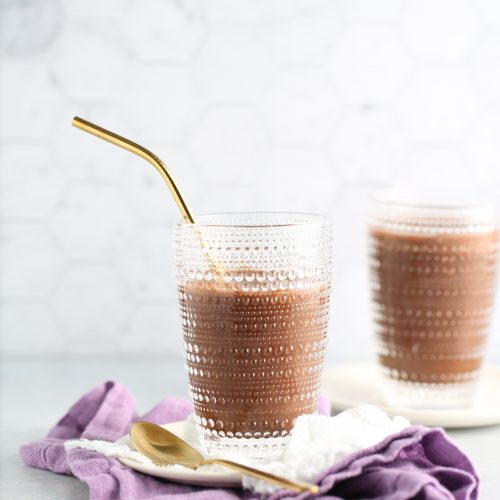 A textured glass of chocolate milk with a gold metal straw sits on a purple cloth, beside a gold spoon and a scoop of powdered ingredient—an inviting setup for any vegan frappuccino recipe, all set against a light hexagonal tile backdrop.