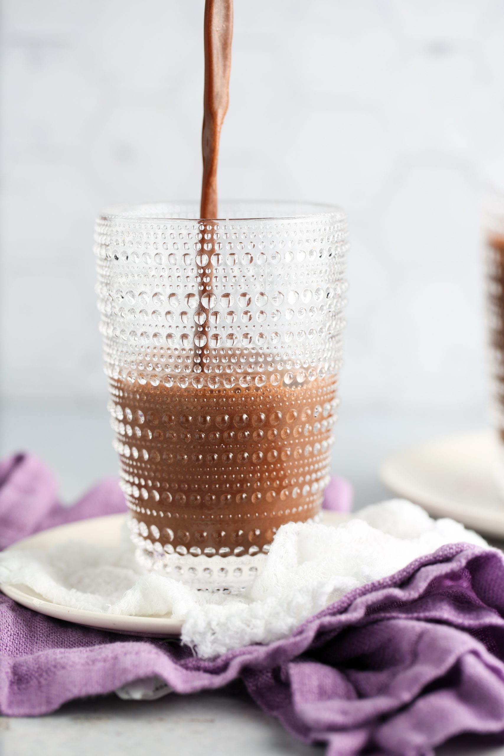 A thick chocolate drink, inspired by a vegan frappuccino recipe, is poured into a textured glass. The glass rests on a white plate with a white and purple cloth casually draped underneath, set against a soft, light background.