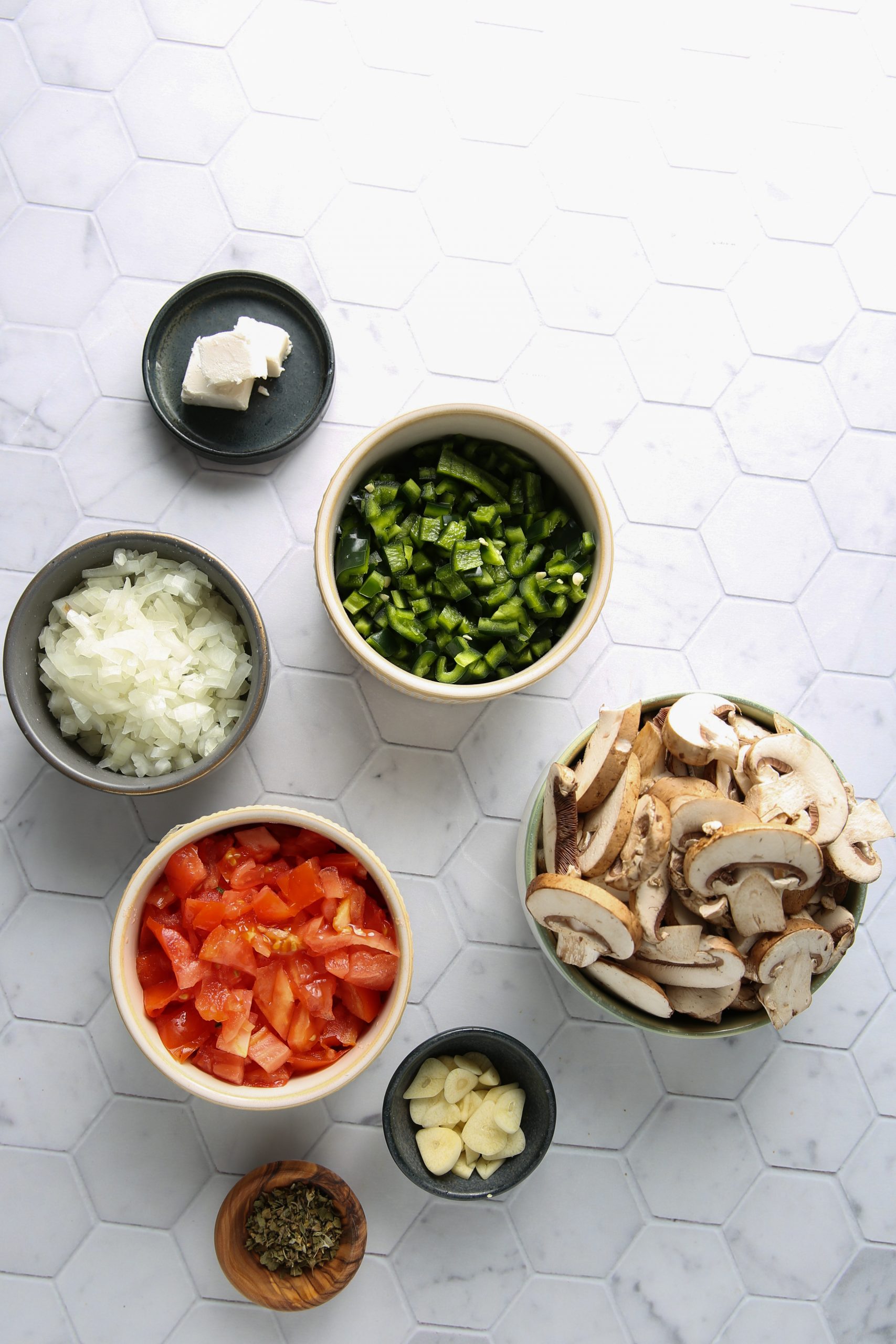Una vista de arriba abajo muestra cuencos con verduras picadas -pimiento verde, cebolla, tomate, champiñones y ajo- junto a queso y hierbas secas. Ingredientes perfectos para preparar tacos gobernador veganos sobre una superficie de azulejos hexagonales blancos.