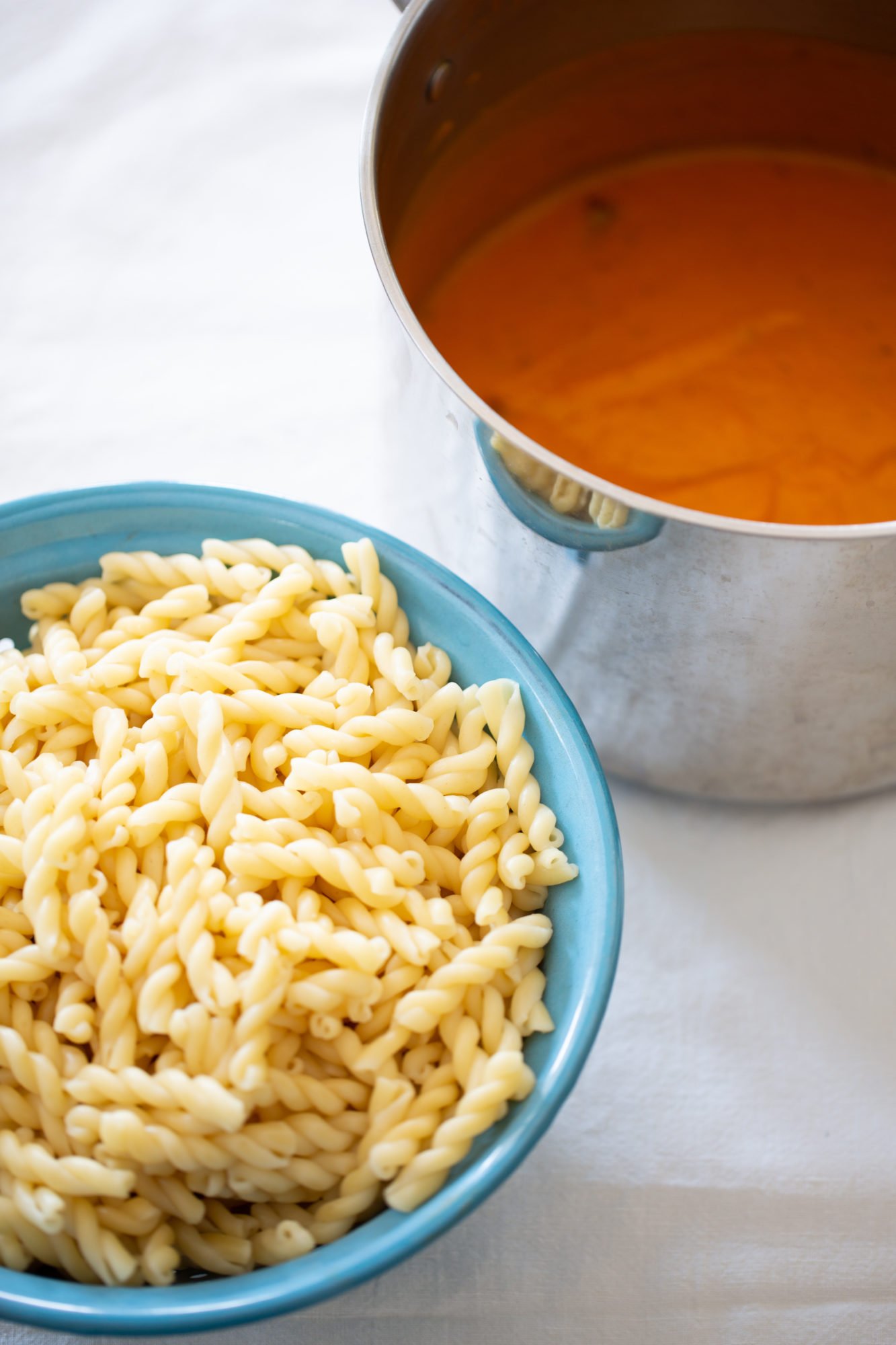 A blue bowl filled with cooked gemelli pasta sits beside a silver pot containing orange-colored sauce. Both items rest on a white surface, suggesting meal preparation in progress.