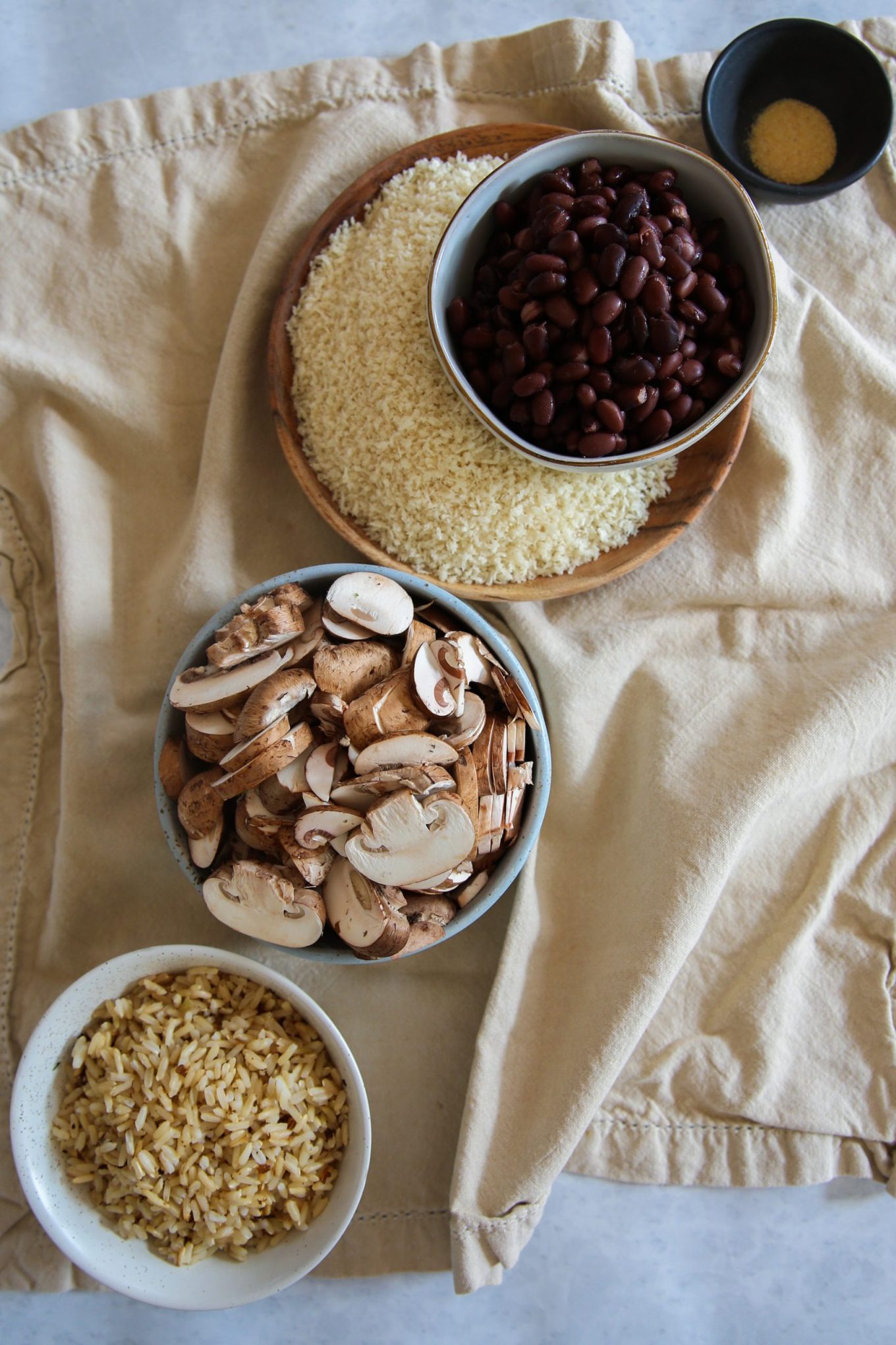 Four bowls sit on a beige cloth. The bowls hold, clockwise from top: brown rice and kidney beans for vegan albondigas, granulated garlic, sliced mushrooms, and cooked brown rice. The casual setting hints at a cozy meal with chipotle flavors.