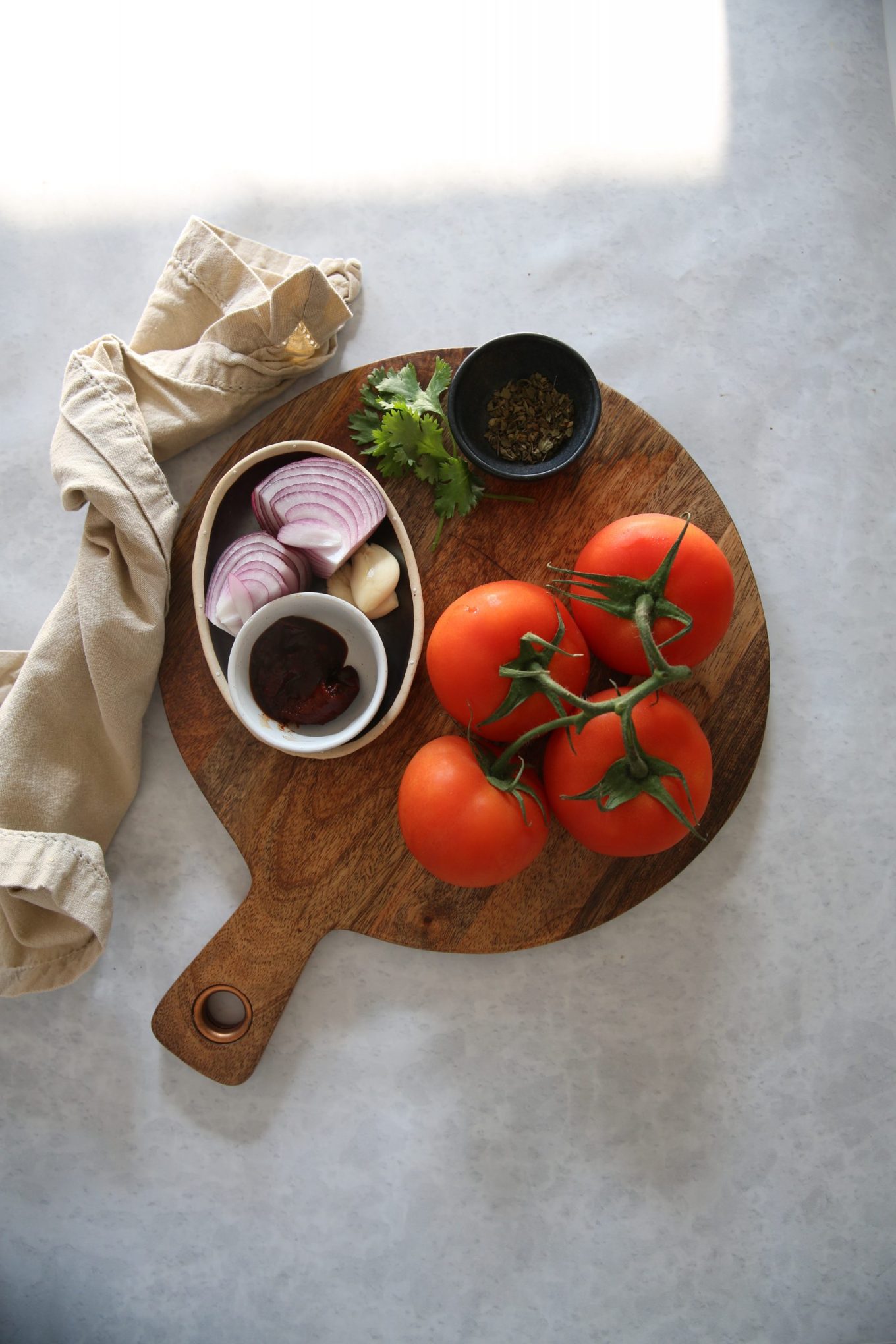 A round wooden cutting board holds vine tomatoes, a small bowl with sliced red onion and garlic, chipotle, a dish with herbs, sun-dried tomatoes, and a sprig of cilantro. A beige cloth napkin rests on the side atop a light stone surface.