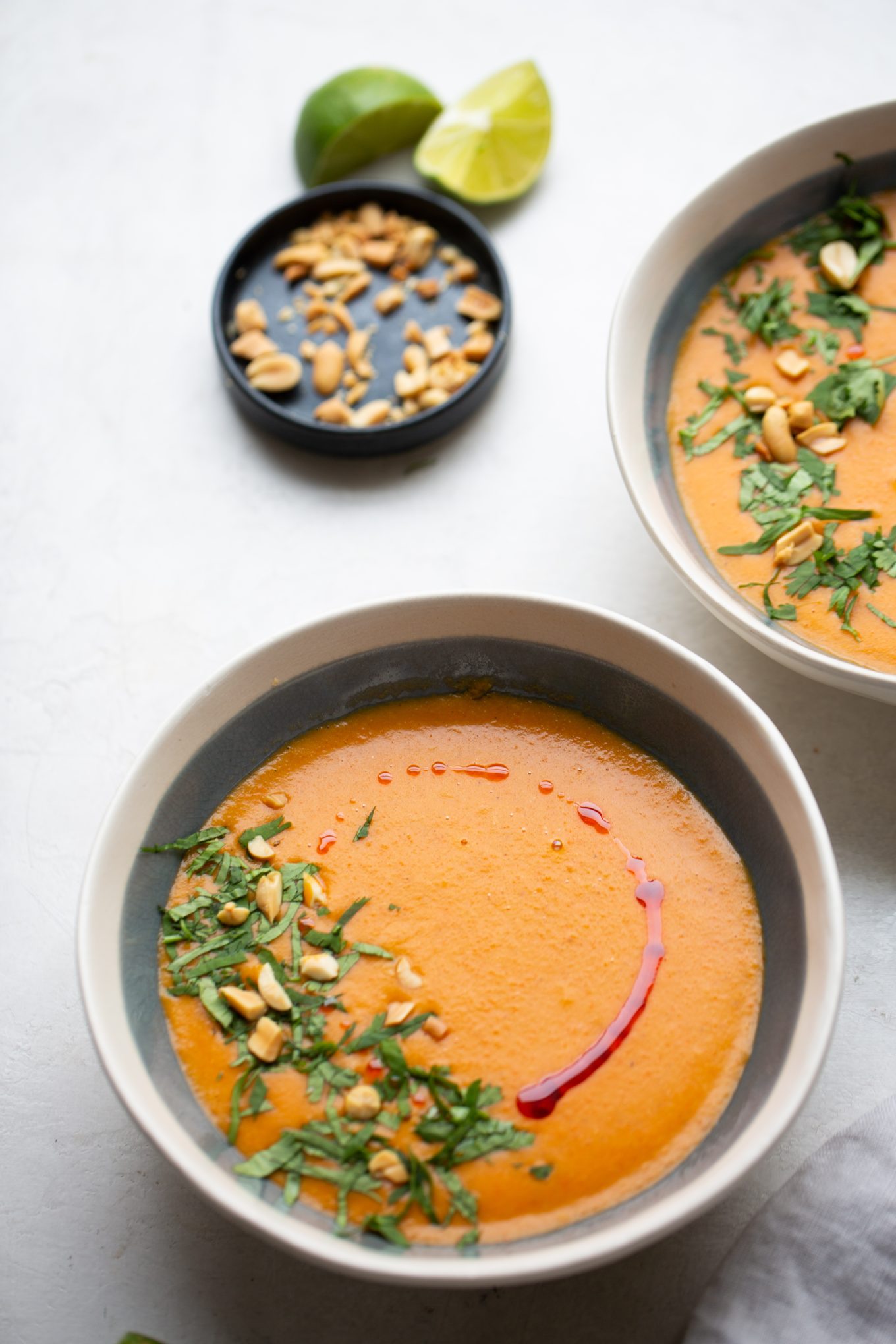 Two bowls of creamy orange vegan red lentil soup are elegantly garnished with chopped herbs, peanuts, and a drizzle of red oil on a light surface. In the background, a black dish holds extra peanuts alongside two lime halves, adding a vibrant touch of green to the composition.