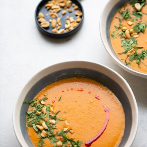 Two bowls of creamy orange vegan red lentil soup are elegantly garnished with chopped herbs, peanuts, and a drizzle of red oil on a light surface. In the background, a black dish holds extra peanuts alongside two lime halves, adding a vibrant touch of green to the composition.