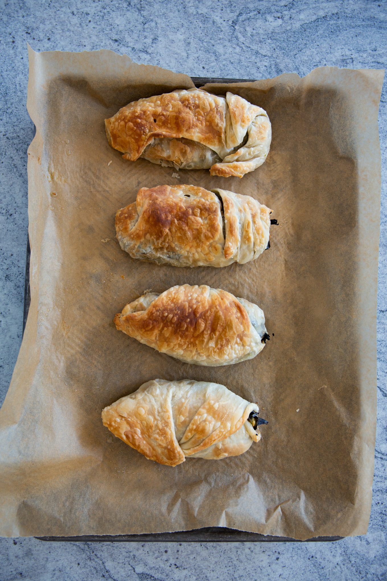 Four golden-brown pastries are lined on a parchment paper-covered baking sheet. Their flaky, puff pastry appearance and uneven, curved shapes suggest they might be homemade puff pastry-wrapped stuffed peppers. The textured grey background enhances the warm tones of the pastries.