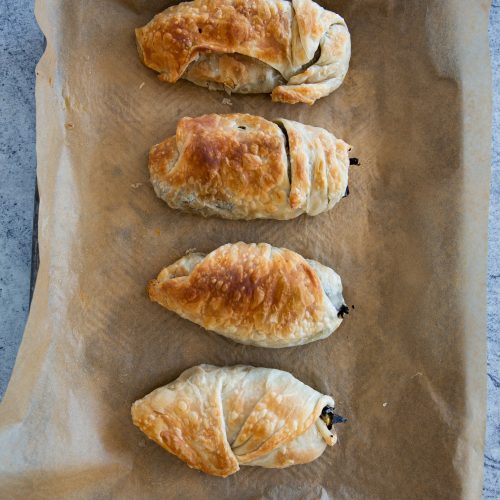 Four golden-brown pastries are lined on a parchment paper-covered baking sheet. Their flaky, puff pastry appearance and uneven, curved shapes suggest they might be homemade puff pastry-wrapped stuffed peppers. The textured grey background enhances the warm tones of the pastries.